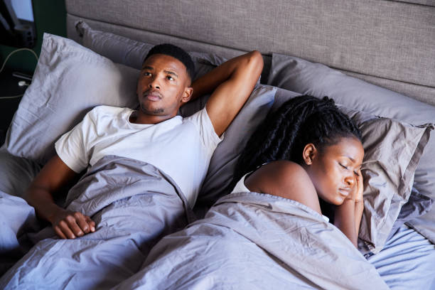 Young African man looking stressed out while lying awake next to his sleeping wife in their bed in the morning